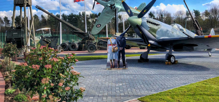 Photo of the entrance of Eden Camp, showing the paved forecourt with a replica full-size Spitfire. It is a bright sunny day with some clouds in the sky and flowers blooming.