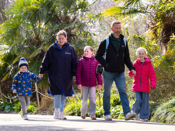 A family walk hand in hand along a paved path through a lush, leafy zoo trail with plants and trees behind them.
