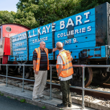 Two volunteers wearing high vis orange vests by a blue railway cart reading 'John L.L. Kay Bart, Denby Grange Collieres No. 9'