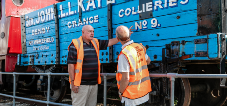 Two volunteers wearing high vis orange vests by a blue railway cart reading 'John L.L. Kay Bart, Denby Grange Collieres No. 9'