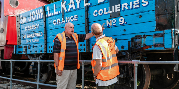 Two volunteers wearing high vis orange vests by a blue railway cart reading 'John L.L. Kay Bart, Denby Grange Collieres No. 9'