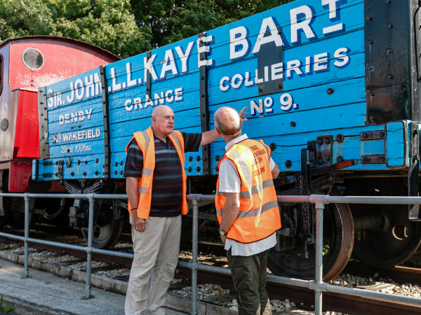 Two volunteers wearing high vis orange vests by a blue railway cart reading 'John L.L. Kay Bart, Denby Grange Collieres No. 9'