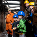 Two young boys with hard hats and lamps eagerly waiting with a Mine Guide and family to go Underground