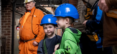 Two young boys with hard hats and lamps eagerly waiting with a Mine Guide and family to go Underground