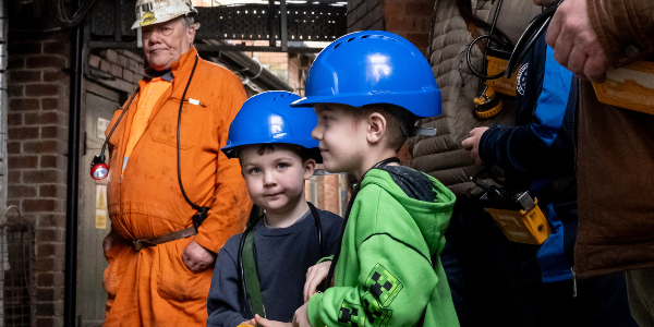 Two young boys with hard hats and lamps eagerly waiting with a Mine Guide and family to go Underground