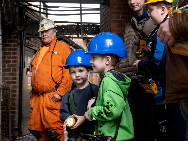 Two young boys with hard hats and lamps eagerly waiting with a Mine Guide and family to go Underground