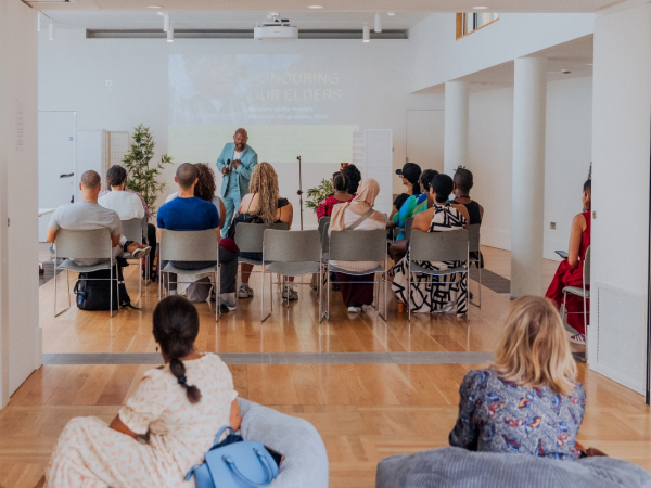 An audience watches a presenter speak, some on beanbags, plants at the sides