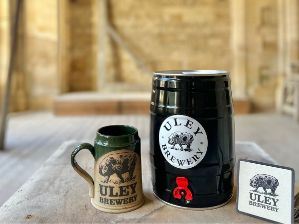 Promotional image showing Uley Brewery products inside Woodchester Mansion. A black mini beer keg with a red tap, a ceramic ale mug, and a brewery coaster sit on a wooden table, with the Mansion’s unfinished stone interior softly blurred in the background