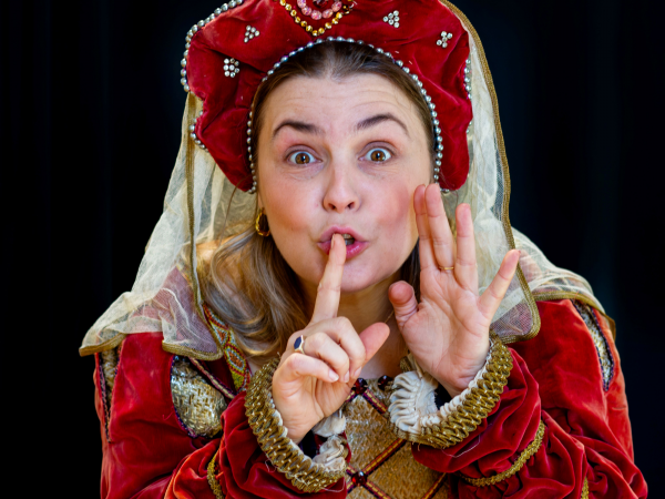 A woman in red and gold medieval style costume holds a finger to her lips, signalling a secret. The background is dark, emphasizing a playful, secretive mood.