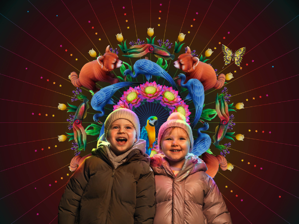 Two warmly dressed children stand in front of a colorful illuminated display featuring animals and flowers.