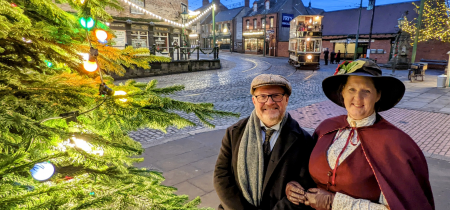 Christmas Evening at Beamish. Edwardian gentleman and lady stand beside a christmas tree with large bauble lights on. In the background is the Edwardian street with a tram waiting at the tram stop.