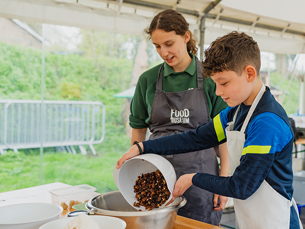A Food Museum employee smiles whilst a child pours a bowl of sultanas into a hot cross bun mixture