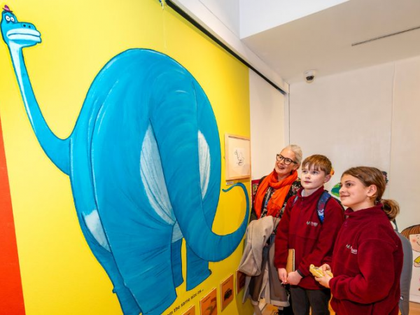 School children and their grown-up on the right look at an impressive blue dinosaur illustration on the yellow wall