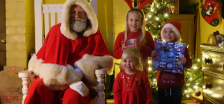 Three children in festive outfits posing with Santa and their presents in Santa's Grotto