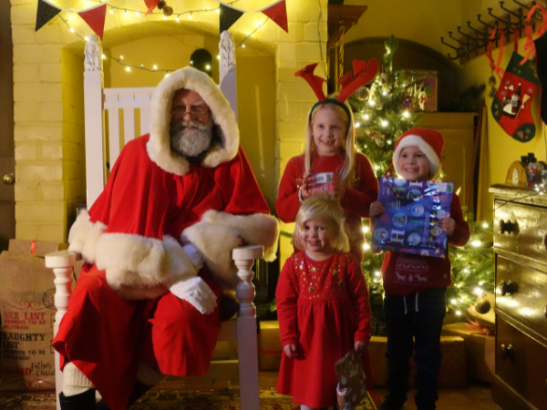 Three children in festive outfits posing with Santa and their presents in Santa's Grotto