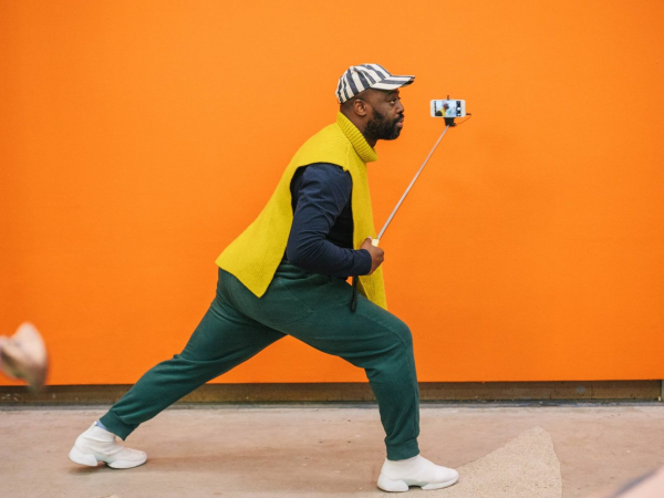 Harold Offeh lunging whilst holding a selfie stick, in front of an orange wall.