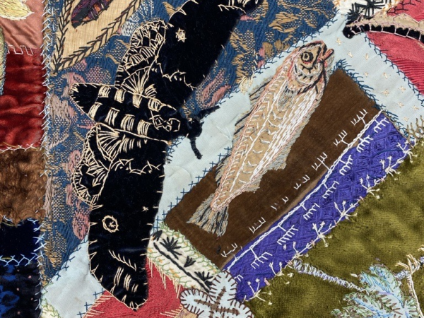 A close-up section of a patchwork quilt with many different random elements including a white bird, a butterfly and abstract patterns, mentions of dates 1892 and 1895.