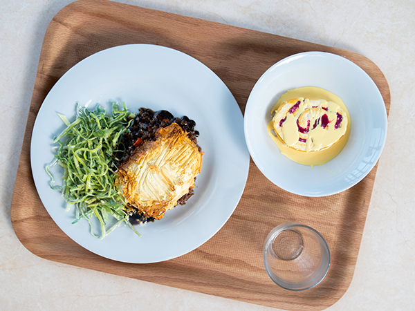 An image of a typical school dinner on a tray, a shepherds pie with a side of greens and a bowl of jam roly poly and custard