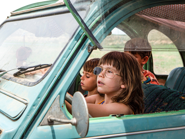 a young girl drives an old car, wearing sunglasses with just one lens. Other young children are behind her.