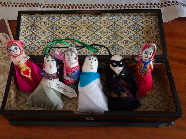 A collection of six handmade dolls displayed in an open wooden case, each embelished with different materials, faces and outfit designs.