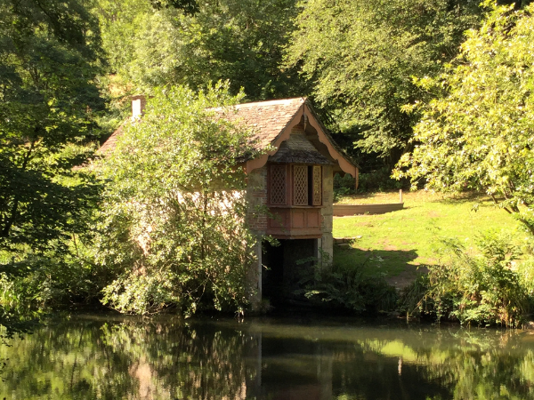 View across a calm lake towards the Boathouse in Woodchester Park. The small Victorian building sits at the water’s edge, partially surrounded by trees and greenery, with sunlight filtering through the woodland and reflecting in the still water below.
