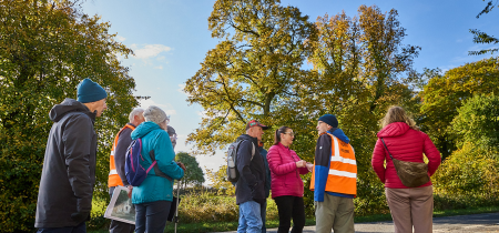 A group of visitors walking up a gradual hill by the water treatment plant by the woodland. In the distance you can see the colliery building.