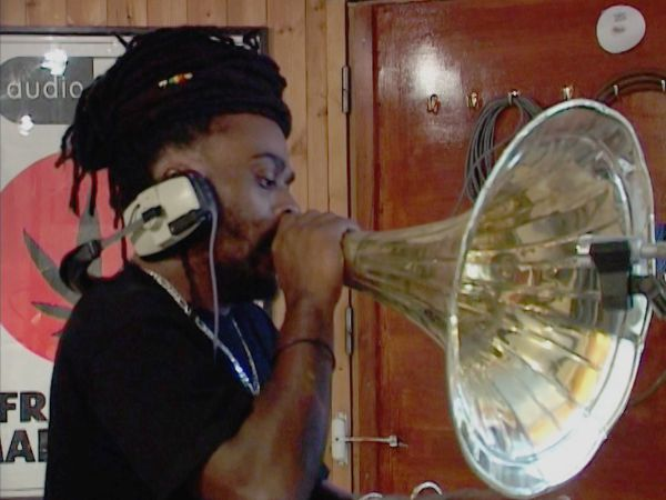 A man with dreadlocks hold a grammaphone horn towards a michrophone.