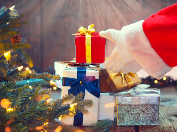 Santa Claus places a red gift box on a stack of presents near a decorated Christmas tree.