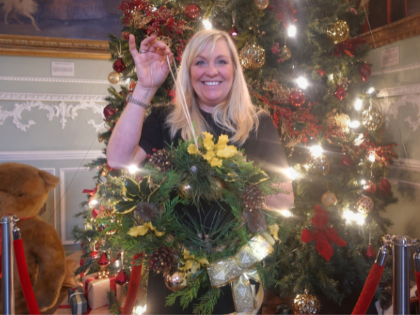 Woman smiling and holding up a Christmas wreath in front of a Christmas tree