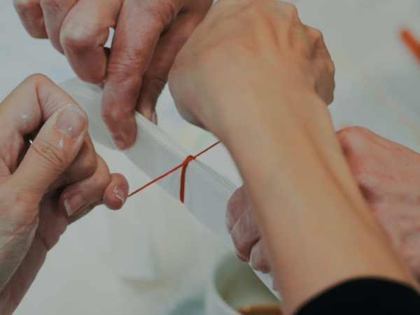 Two pairs of hands, one marked with spots of white paint, collectively tie a red string around layers of a folded strip of paper.