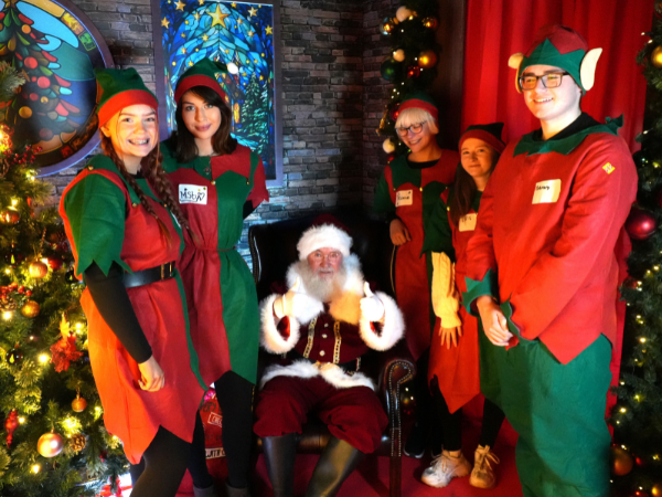 Santa Claus seated in a festive chair, surrounded by five staff members dressed as elves, with decorated Christmas trees, colourful stained-glass-style panels, and warm holiday lighting in the background.