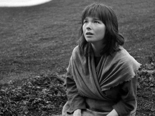 A black and white film still, of a seated girl looking up against a barren hillside.