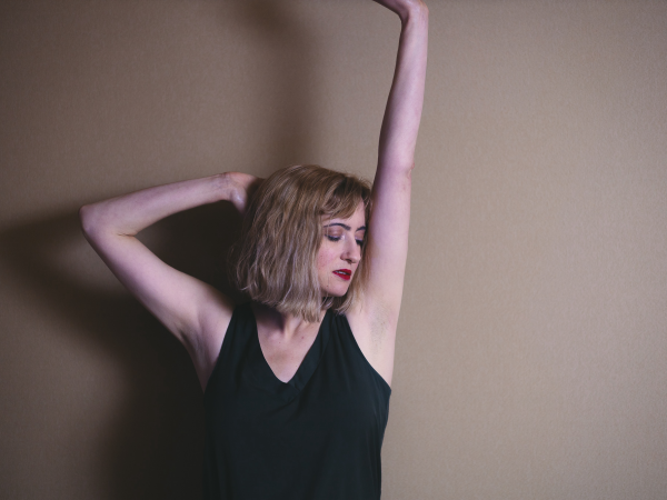 A woman white short blonde hair with her arms stretched above her head, her head tilted to one side.