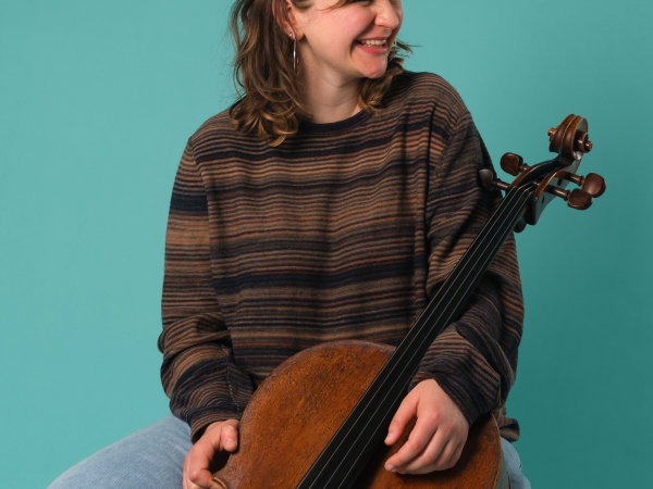 A woman in a knitted jumper smiling, and holding a cello.