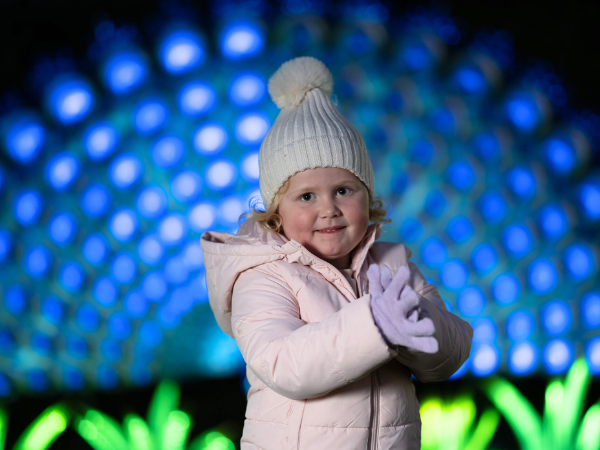 A child in a white knit hat and pink jacket stands in front of a glowing blue and green illuminated display, evoking a magical winter atmosphere.