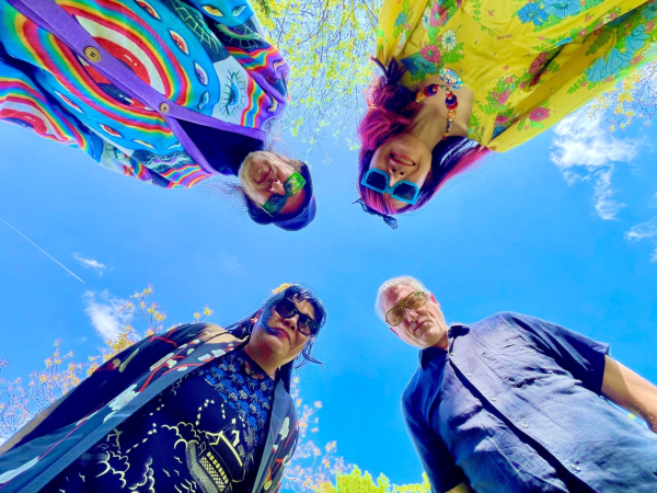 4 band members in colourful outfits stand around with the camera pointing up to the sky.