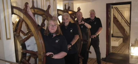 The four members of Spinnaker Shanty standing in a vertical row. In the front is the female member of the group. They are onboard HMS victory holding the ships wheel.