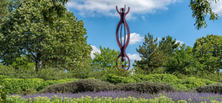 The sculpture 'Chain Reaction'. In the foreground there are purple flowers. The sculpture is framed by trees.