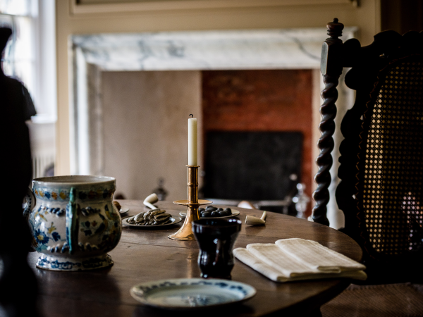A table from a 1695 Parlour, with posset pot, candle stick and dried food, with a darkened foreground with two chairs.