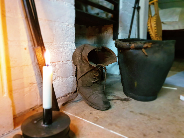 A worn rough leather boot is propped against a brick basement wall, lit by a candle.