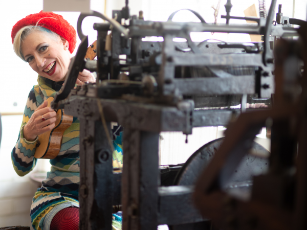 Cabaret artist Tricity Vogue sitting at an antique knitting frame. Wearing a stripy knitted dress, she has a broad smile and is playing the ukulele.