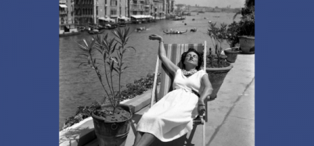 Black and white photograph of Peggy Guggenheim sat laid back in a deck chair on a patio in front of the grand canal, Venice.