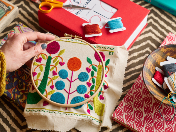 A hand holds an embroidery work in process amongst a backdrop of brightly coloured prints and materials.