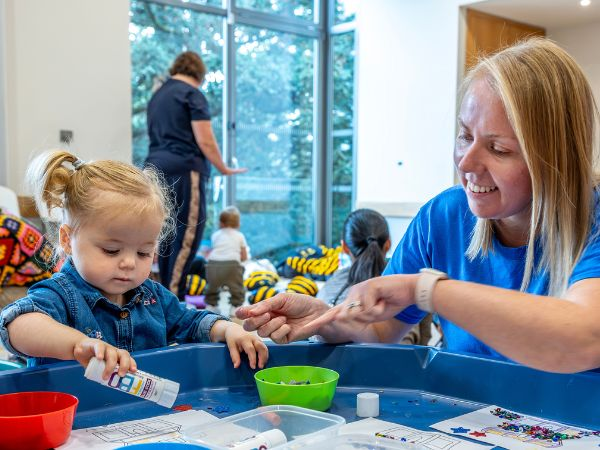 A toddler on the left joins session leader Laura on the right in a crafts activity at a blue table