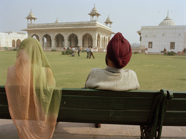 Two people sat on a park bench overlooking a grassy square in Delhi. The person on the right is wearing a translucent orange and green sari, and the person on the right is wearing a knitted vest and a red turban.