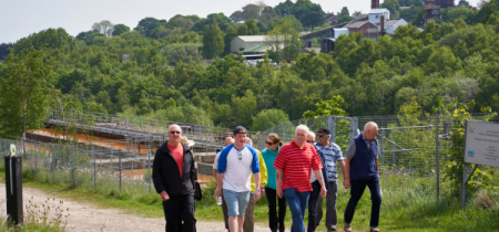 A group of visitors walking up a gradual hill by the water treatment plant by the woodland. In the distance you can see the colliery building.