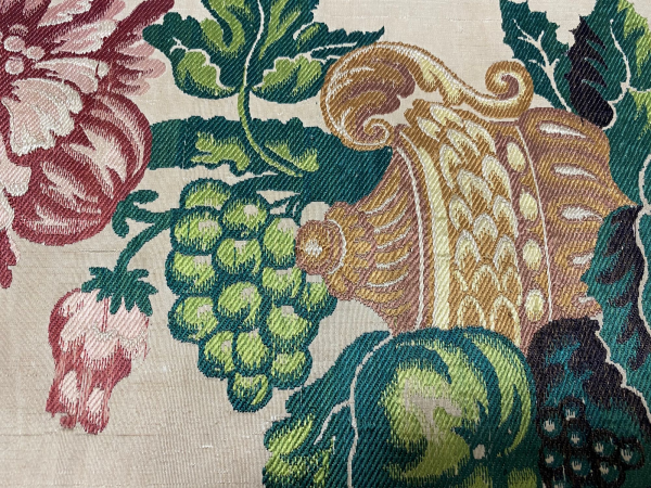 Detailed close up of a silk textile, with a orange pot, green leaves and grapes and a red flower.
