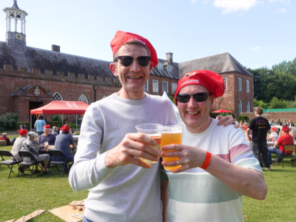 Two men wearing Chouffe beer hats and sunglasses hold beer and smile outside Hartlebury Castle