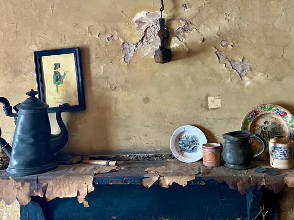 plates cups and a jug sit on a rugged mantlepiece in front of a peeling tan-coloured wall