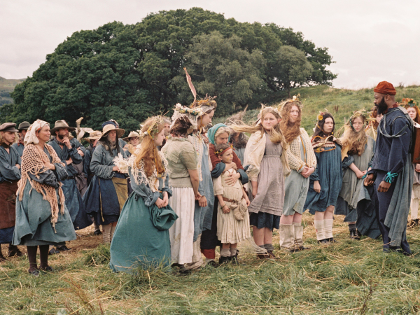 Film still, showing a large group of people in dresses and hats stood in a hilly field with trees in the background.